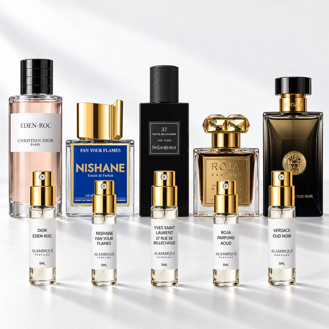 Discovery set Alambique: DIOR EDEN-ROC, NISHANE FAN YOUR FLAMES, YVES SAINT LAURENT 37 RUE DE BELLECHASSE, ROJA PARFUMS AOUD, VERSACE OUD NOIR. Decants con etiquetas blancas y atomizadores dorados junto a las botellas de referencia.